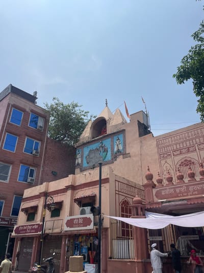 The image shows a street scene in Amritsar, India, with a focus on a building adorned with religious motifs and signage in Punjabi. The building, likely a temple or gurdwara, has a pale pink facade with intricate red decorative patterns. Above the entrance, there is a large sign with Punjabi script. A white canopy is extended over the entrance, and several people are visible in the foreground, some in traditional attire. One person, dressed in white with a white turban, appears to be interacting with someone on the other side of a counter. Adjacent to this building is a taller, modern building with blue-tinted windows and air conditioning units. The sky is clear blue, and a tree with green foliage is visible to the right. Signs in Punjabi for "Veere Cafe" and "N Railway Ticket Office" are present on the lower part of the building. The overall scene depicts a bustling urban environment with a strong cultural and religious presence.