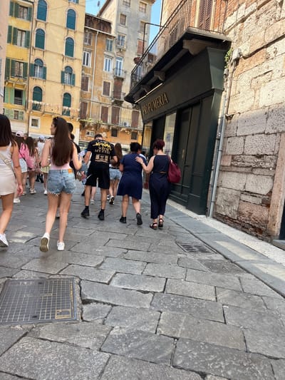 The image shows a group of people walking on a cobblestone street in Verona, Italy. The buildings are old, with colorful facades and many windows with closed shutters. A woman with a burgundy backpack is standing in front of a shop, which has a dark green awning with the word "PROFUMERIA" written on it. The people are walking in the same direction. The weather is sunny and the light is bright.