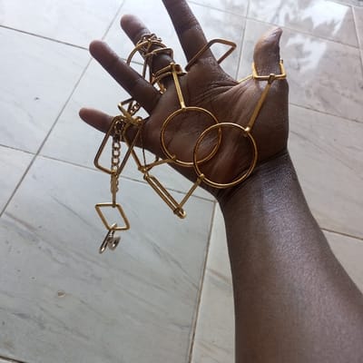 A human left forearm and hand, exhibiting dark brown skin, is presented in a high-angle, close-up view against a light grey tiled surface in Mahiakalo ward, Kenya. The hand is positioned palm-up, with fingers slightly curved, supporting a collection of interlocking metallic objects. These objects are primarily gold-toned, forming a complex, open-ended chain-like assembly of geometric shapes, including prominent circular rings and various rectangular frames. Smaller, finer gold chain segments connect some of these elements, and a single silver-colored clasp or small key is suspended from one end of the assembly. The tiled background displays visible grout lines.