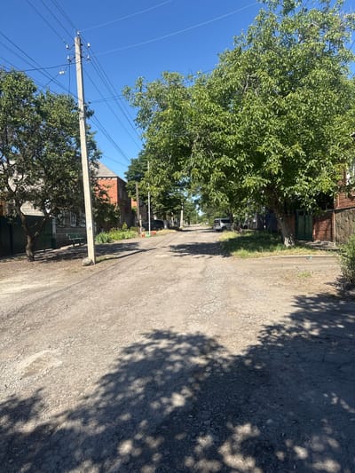 This is a vertical, eye-level, outdoor shot of a gravel road in a residential area. The sun is shining brightly, casting distinct shadows from trees onto the road and the surrounding buildings. The sky is a clear, vibrant blue.
On the left side of the road, a large, leafy green tree dominates the foreground, its branches reaching upwards. A utility pole with numerous wires stretches vertically beside the tree, extending towards the sky. Behind the tree, glimpses of a brick building with green doors and windows can be seen. Further down the road, more trees and buildings are visible, suggesting a rural or suburban setting. A dark-colored car is parked on the side of the road in the distance.
The road itself is rough and appears to be made of gravel and asphalt. The overall impression is a warm, sunny day in a quiet, possibly older neighborhood. The image does not contain any discernible text. The scene likely depicts a typical day in a residential area.