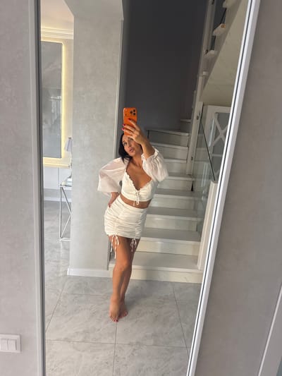 In Malinovka, Russia, a woman stands in a modern interior, taking a selfie in a full-length mirror. She is wearing a white ruched crop top with drawstring details and matching ruched mini-skirt with drawstring ties. Her hair is dark and pulled back, and she is wearing bright red nail polish. Her body is positioned at an angle, with one hand on her hip. Her reflection is visible, showing a contemporary staircase with white steps and glass railings behind her. The flooring is light-colored tile with a marble pattern. To the left of the mirror, another reflection shows a hallway with a console table and a lamp. The wall appears to be a textured gray.