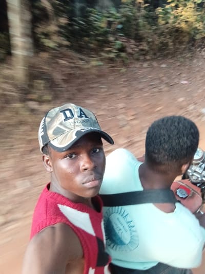 Two people ride a motorcycle down a dirt road. The person in the foreground is wearing a camouflage cap with the word "Dallas" printed on it, a red and black sleeveless top. The person behind him wears a light blue t-shirt. The road they travel on is surrounded by trees. The image is slightly blurry, suggesting movement.