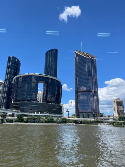 The image captures a vibrant daytime scene of modern architecture along a river in Brisbane City, Australia. The perspective is from the water, looking towards the riverbank.

On the left, several prominent skyscrapers dominate the skyline. The tallest, a sleek, dark-glass tower with a distinctive curved top, stands out. Below it, a complex of contemporary buildings features a unique multi-level design with dark glass facades and some greenery on terraces. One building within this complex clearly displays a white logo that reads "THE STAR". Further down the river, another dark-glass high-rise is visible, along with a lighter-colored, older-style building in the background. A white construction crane is visible between the buildings, indicating ongoing development.

The river, likely the Brisbane River, occupies the right side of the frame. Its surface is rippled and reflects the bright sunlight, appearing a warm, muddy brown color. Along the riverbank, trees and an elevated pedestrian walkway or bridge structure can be seen.

The sky is a clear, bright blue with scattered white cumulus clouds. Several faint, elongated, parallel light streaks are visible in the upper part of the sky, likely reflections or lens artifacts rather than actual objects.

The overall impression is one of a bustling, modern city on a sunny day. No people or specific activities are distinctly visible, but the urban landscape suggests a lively environment. The time of day is clearly daytime, given the strong illumination and blue sky.