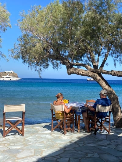The image depicts a sunlit coastal scene in Φάρος, Greece, focusing on a patio area bordering the sea. In the foreground, an irregular light-toned flagstone patio is partially shaded by a large, mature tree with a prominent, gnarled trunk extending from the right side of the frame. An empty wooden director's chair with off-white canvas is positioned on the left side of the patio. In the midground, a square wooden table, covered with a light-colored cloth accented by blue and white patterned fabric, is situated at the patio's edge, directly overlooking calm, clear turquoise waters. Two individuals are seated at the table, their backs partially visible to the viewer. The individual on the left, a female, wears a yellow halter-neck top and a patterned yellow and white sarong. The individual on the right, a male, wears a dark blue short-sleeved shirt and a light-colored baseball cap. Both individuals are looking down at white papers or menus on the table. In the background, the blue sea extends to the horizon. A large white multi-deck superyacht is anchored in the left mid-distance, positioned off a small, white, traditional Greek building situated atop a coastal headland. A smaller, less distinct vessel is faintly visible further to the right on the water. Several small yellow buoys are dispersed across the water's surface.
