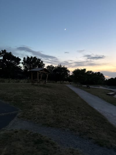 The image depicts a serene park scene at dusk in Saint-Orens-de-Gameville, France. A wooden gazebo with a picnic table and benches is centrally located on a grassy hill. To the right, a paved pathway winds through the park, bordered by trees and benches. In the distance, the sky displays a gradient of colors from blue to yellow, with a crescent moon and a faint star visible. The overall atmosphere is peaceful and natural.