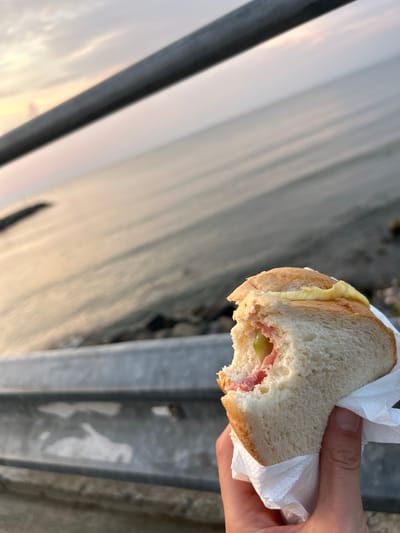 A hand holds a partially eaten sandwich wrapped in white paper, with a bite taken out of it. The sandwich appears to contain pink ham and green slices. The background is a blurry body of water and sky, possibly at sunset, with a metal railing in the foreground. The scene is set in Ravda, Bulgaria.