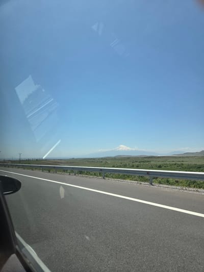 The image presents a bright, clear daytime view captured from inside a vehicle, looking out across a rural or semi-rural landscape. The scene is dominated by a vast blue sky in the upper portion. In the middle ground, green fields and rolling terrain extend towards the horizon. In the distance, a majestic snow-capped mountain, likely Mount Ararat given the Kosh, Armenia context, is visible, its peak appearing white against the lighter blue, hazy distant sky.
A grey metal guardrail runs along the right side of the frame, separating the green roadside from the paved asphalt road. The road features a distinct white solid line. In the bottom right corner, a portion of the vehicle's dark interior, possibly a dashboard or door frame, and a wheel arch are visible. The left side of the image is affected by significant reflections on the vehicle's window, showing abstract light-colored shapes, including parallel lines and angular forms, obscuring parts of the exterior view. The weather appears sunny and clear. There is no visible text in the image.