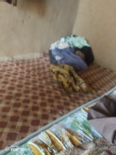 In Gulak, Nigeria, an indoor scene captures a bed covered with a brown and maroon patterned sheet. A pile of clothing, including items in light blue, green, and black, rests on the right side of the bed. Another garment, with a brown and yellow pattern, is draped across the bed. The walls appear to be a light tan color. A portion of a mobile device's watermark, "TECNO POP 10," is visible in the lower left corner of the image.