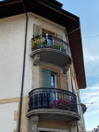 This is an eye-level, medium shot of a corner of a stucco-clad building in Milvignes, Switzerland, featuring two balconies. The building has a dark brown, sloped roof with exposed wooden beams. The top balcony is adorned with yellow flowers and greenery in pots, with a dark wrought-iron railing. A window with green shutters is visible behind the railing. The lower balcony also has a dark wrought-iron railing and contains pink flowers in pots. A window with green shutters is also visible behind this railing. Both balconies are supported by stone corbels. A black drainpipe runs vertically along the side of the building. The sky is a clear blue with some wispy clouds. The overall scene depicts a charming residential building exterior.