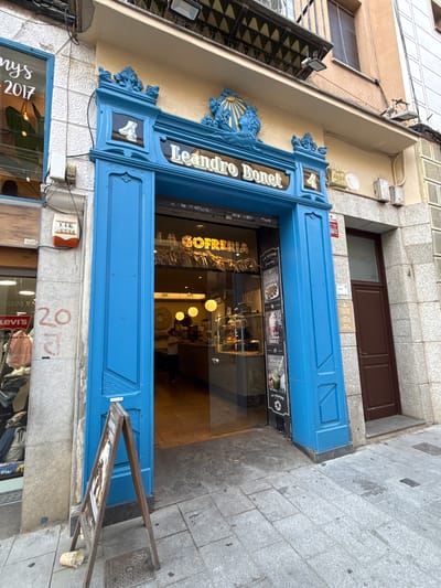 The image shows the exterior of a shop with a bright blue entrance. Above the entrance, the name "Leandro Bonet" is displayed in white lettering. Below this, the words "LA GOFRERIA" are visible in a yellow, stylized font, indicating it is a waffle shop. A sandwich board sign stands on the sidewalk in front of the entrance. To the left of the blue entrance, a store window displays clothing and the year "2017". To the right of the blue entrance is a brown door. The shop is located on a paved street. A person is visible inside the shop, standing behind a counter. The building facade is painted a light yellow and textured grey.