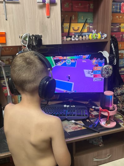The image shows a young person, from behind, wearing headphones and playing a video game on a computer. The setting appears to be a room with shelves filled with vintage-style luggage. A pink microphone is mounted on a stand next to the computer monitor, which displays a game with a purple background and various interface elements. On the desk, there's a keyboard with blue-backlit keys and other gaming accessories. Several small figurines are arranged on the shelf above the monitor, and a pair of sunglasses rests on the desk in the foreground. The lighting suggests it's daytime, indoors. The person's posture and the presence of gaming equipment indicate they are engaged in playing the game. The background shelves with luggage suggest a personal space, possibly a bedroom or study. There is some text visible on the monitor interface, including "Spotify," "Free," and "Hook." There is also text on the figurines on the shelf and on some items on the desk.