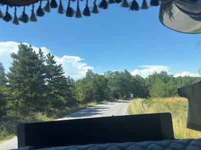 The image captures a view from inside a vehicle traveling on a paved road through a forested area with a bright blue sky and scattered clouds. In the foreground, a portion of the vehicle's dashboard is visible, along with decorative tassels hanging from the top of the windshield. The road ahead curves to the right, flanked by tall green trees and dry grass on either side. Sunlight casts shadows on the road. In the distance, a white sign is visible on the right side of the road. The scene depicts a rural or semi-rural environment, possibly in Hliabovo, Bulgaria, given the context provided.