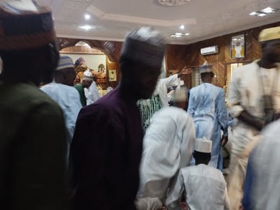 The image shows a gathering of people in a room with a wood-paneled ceiling. The room is lit by overhead lights, and there is an air conditioner on the wall. The people are all wearing traditional clothing, including white robes and caps. Some are wearing long-sleeved shirts. The man in the center of the image is the most prominent figure, wearing a purple long-sleeved shirt and a white cap. He is standing, looking towards the left of the frame. There is a white-robed man with a white cap standing to the right of him. There is a large group of people in the background, many are standing and some are sitting. The coordinates provided show the location to be in Kaltungo, Nigeria. The scene is taking place indoors, in a room that appears to be a gathering space.