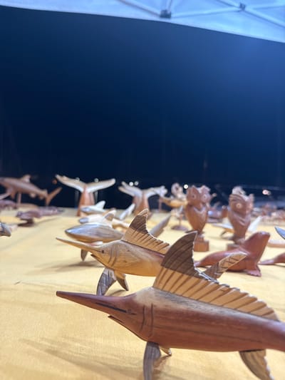 A close-up, low-angle shot captures a collection of carved wooden marine animals and owls displayed on a textured, golden-yellow surface, likely a market stall in Port d'Alcúdia, Spain. The foreground is dominated by two carved marlin figures, their distinctive bills and striped fins rendered with detail. Behind them, a variety of other wooden carvings are visible, including several more fish, stylized whale tails, and a pair of carved owls. The background is dark and out of focus, with hints of a white tent structure above, suggesting an outdoor setting, possibly a night market or a stall set up under shelter. The lighting appears to be artificial, creating soft highlights and shadows on the carved objects.