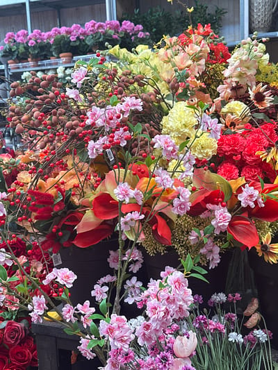 The image displays a close-up of a vibrant and abundant arrangement of artificial flowers, likely in a retail or decorative setting. The foreground is dominated by clusters of pink and red berries on thin brown stems, intertwined with various flowers in shades of pink, yellow, red, and orange. Notable floral elements include dense red carnation-like blooms, delicate pink blossoms, and larger, trumpet-shaped red and green leaves. Some branches with small, round, reddish-brown fruits are also visible. In the background, rows of potted pink flowers are seen on shelves, suggesting a garden center or florist shop. The overall impression is one of rich texture and color, characteristic of a floral display. There is no discernible indication of the specific location in Tuk, Netherlands, within the visual information provided.