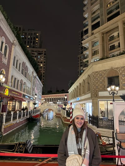 The image displays a nighttime scene within a Venetian-themed commercial and residential complex, identified as being located in Esenler, Turkey. In the immediate foreground, a young adult female stands centered, facing and smiling directly at the camera. She wears a cream-colored knit beanie with a pom-pom, a grey and striped scarf, a dark brown hooded sweatshirt, and carries a quilted beige shoulder bag.

Behind the subject, a narrow, illuminated canal with green water extends into the background. A white arched bridge spans the canal in the mid-ground. Two dark-colored gondola-style boats are present in the canal, one partially visible in the lower left foreground and another further back near the bridge.

Buildings with multi-story, Italianate-style architecture line both sides of the canal, featuring arched windows, detailed facades, and illuminated storefronts displaying various business signs, including "KİĞILI" and "Super Viga." Pedestrian walkways run alongside the canal, populated by several individuals. Tall, modern apartment buildings rise in the distant background, their illuminated windows contrasting with the dark night sky above the themed complex. Artificial light sources, including ornate street lamps, illuminate the scene and reflect on the canal's surface.