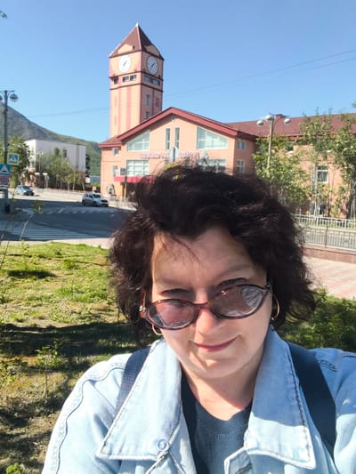 In this selfie, a woman with dark, curly hair and sunglasses stands in front of a building with a prominent clock tower, set against a clear blue sky. The building, painted in light orange and accented with red, appears to be a public structure, possibly a train station or administrative building. The clock tower is tall and slender, featuring a brown roof and four clock faces. In the background, a paved road with a crosswalk is visible, with two cars driving on it. A mountain range or large hill is discernible behind the buildings, suggesting an urban setting within a mountainous region. The woman is wearing a light blue denim jacket over a dark top and a backpack. The foreground features green grass and some foliage. The overall scene captures a sunny day in the city of Kirovsk, Russia, specifically in front of the Kirovsk Railway Station.