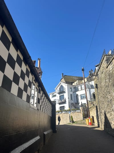 An upward-angled view captures a narrow street or alleyway in Knaresborough, United Kingdom, under a clear, bright blue sky. On the left, a building presents a facade with a lower section painted dark grey or black, topped by a prominent black and white checkerboard pattern extending to the upper edge of the frame. A single rectangular window with reflective panes is visible within this checkered section. Above the building, a light brown chimney with a metal cap projects against the sky.

On the right, a weathered stone wall, likely a retaining wall, runs parallel to the left structure, exhibiting irregular masonry and casting a strong shadow onto the paved ground. The top of this stone wall is adorned with decorative black metal railings.

In the mid-ground, a lone individual, dressed in dark clothing, walks away from the viewer towards a cluster of white multi-story buildings. These buildings feature multiple windows and black-railed balconies. One building roof is visible with light brown tiles. A wooden utility pole, supporting several black wires, stands prominently in the center-right mid-ground. Near the utility pole, a yellow and red traffic cone rests on the pavement. A black street lamp post and a small green potted conifer are also visible near the base of the white buildings. The ground surface is dark and untextured.