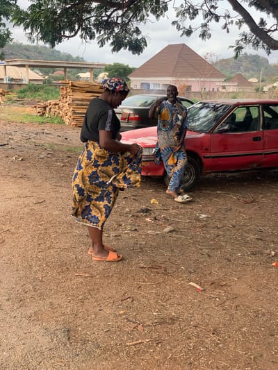 The image shows two people standing near a red car in Kabba, Nigeria. The ground is dirt and gravel. In the foreground, a woman wearing a black t-shirt and a blue and gold patterned skirt is standing. She is also wearing orange sandals and a patterned headscarf. Behind her, there is a stack of lumber. A man wearing a blue and yellow patterned shirt is leaning against the car, next to the woman. A house with a pyramidal roof is visible in the background, as are some trees and a distant hillside. The sky is overcast.