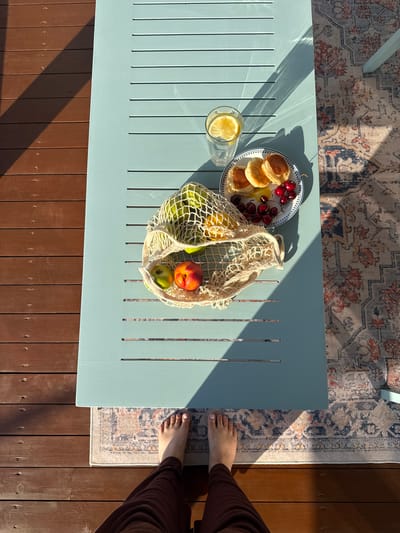 In this overhead shot of a light blue wooden table, a spread of breakfast items and fruit is bathed in warm sunlight, suggesting it is daytime. On the table, a plate holds several small, golden pancakes and a scattering of bright red cherries. Adjacent to the plate is a clear glass filled with water and a slice of lemon. A woven net bag rests near the plate, filled with green apples, a nectarine, and possibly other fruits. The table is situated outdoors, as indicated by the shadow of a rug and the presence of wooden decking beneath it. A patterned rug, partially visible, lies on the decking, and a person's bare feet and brown trousers are visible on the right edge of the frame, suggesting they are sitting at the table. The overall mood is relaxed and pleasant, with the natural light creating a serene atmosphere. The image appears to have been taken in Starokorsunskaya, Russia.