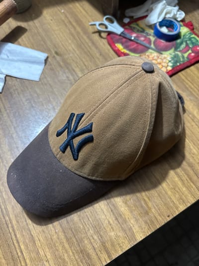 A close-up, eye-level, medium shot captures a tan baseball cap with a dark brim, emblazoned with a black "NK" logo, resting on a wooden table. The cap is positioned slightly off-center, with its front facing towards the viewer. The brim is dark brown, and the main body of the cap is a lighter tan color. The "NK" logo is a stylized black embroidery. A dark gray button sits atop the crown of the cap. The wooden table has a visible grain pattern. In the background, out of focus, are a white folded cloth, a pair of scissors, a roll of blue tape, and a colorful mat with fruit designs. The scene is well-lit, casting shadows from the cap onto the table. This image is not located in Pîrlița, Moldova.