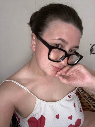 The image is a close-up, waist-up selfie of a young woman wearing black, rectangular glasses and a white camisole adorned with red hearts. She has her hair pulled back into a bun and is resting her right hand on her chin, looking towards the camera. The background appears to be a light-colored wall with some faint markings on the right side, possibly a drawing. The lighting suggests an indoor setting. There is no discernible information in the image that would indicate a specific location within the city of Chaikovsky, Russia.