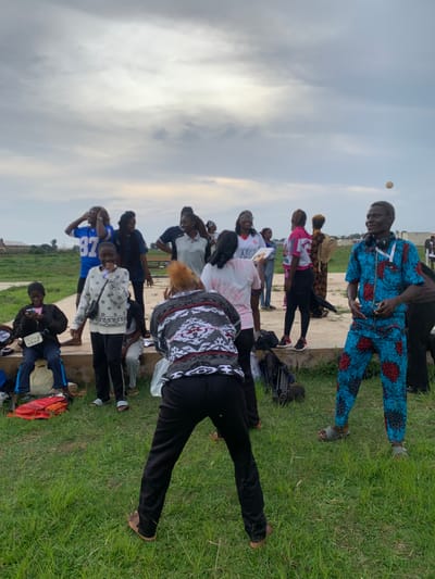 This is a photograph of a group of people gathered outdoors in Gwafan, Nigeria. The image is oriented vertically, with the sky filling the upper portion and a grassy area and a paved surface occupying the lower section. Several people are visible, some standing and some appearing to be seated or in motion.
One person in the foreground is wearing a blue and red patterned outfit. Another person is wearing a white and black patterned shirt with black pants. Further back, several individuals are dressed in casual clothing, including a blue shirt with the number "87" on it. Some people are holding items, such as a book. The group appears to be engaged in a social gathering or activity.
The sky is overcast, suggesting it may be a cloudy day. The time of day appears to be late afternoon or early evening, based on the lighting conditions. The presence of grass and what looks like a pathway indicates an outdoor setting, possibly a park or an open public space. There is a small, light-colored circular object in the sky above the people, which may be a balloon or a similar item.
Some of the people are looking in different directions, and their expressions are not clearly discernible. The overall mood seems relaxed and informal. There are no obvious signs of distress or urgency. The context of Gwafan, Nigeria, places the scene in a specific geographical and cultural setting.