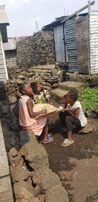 The image captures four young individuals, likely girls, engaged in an outdoor activity in the city of Buzove, Democratic Republic of the Congo. The scene unfolds on a dirt ground surface featuring exposed, dark, porous rock formations and constructed steps leading upwards into the background. Surrounding structures include buildings with corrugated metal walls and roofs, and a prominent wall built from dark, irregular stones. One metal structure on the far right has faded, illegible text on its surface. In the immediate foreground, two girls are seated on large dark rocks, jointly holding a light-colored, rectangular board or tablet with visible markings on its surface, suggesting writing, drawing, or a game. The girl on the left wears a peach-colored sleeveless dress and blue sandals, while the girl on the right wears a white short-sleeved top, dark leggings, and patterned light-colored sandals. A third girl, wearing a green and white patterned dress, is seated behind the left-most girl, looking upwards and to the right, with one hand touching her head. A fourth, younger child is partially visible kneeling or sitting to the right, also observing the board. The environment is illuminated by bright daylight, casting defined shadows.