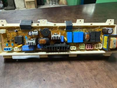 An overhead, eye-level shot shows a dirty, beige plastic-encased circuit board, likely from a washing machine, resting on a brown, wood-grain textured table. The circuit board is populated with various electronic components, including several black relays, blue capacitors, copper-wound transformers, heatsinks, and white connectors. Some components are coated in a yellowish residue. The board is secured to a larger white plastic frame with mounting holes. In the blurry background, dark boots are visible on the left, and a partially obscured green surface is on the right. The scene appears to be an indoor workshop or storage area.