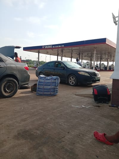 At a SALBAS Oil & Gas station in Galadimawa Village, Nigeria, two dark-colored sedans are parked under a covered canopy. The car on the left has its trunk open, and a person in an orange shirt is standing nearby, appearing to interact with the car's contents. A large, checkered duffel bag rests on the ground between the two vehicles. A spare tire is visible under the rear of the car on the left. The car on the right is closer to the gas pumps, and another person in a white shirt and cap is standing near its driver's side door. A black rolling suitcase with red accents is positioned on the ground in front of the car on the right. The ground is paved with concrete, and some water or liquid stains are present. In the foreground, a person's foot wearing a red slip-on shoe is visible, resting on the ground. The sky is bright and clear.