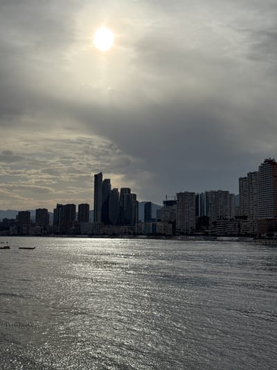 This is a vertical, eye-level shot of a cityscape by the sea. The sun, a bright white orb, is positioned in the upper left quadrant of the image, partially obscured by wispy clouds. The sky is overcast, with the sun's light diffused and creating a bright haze. Below the sky is a dense collection of high-rise buildings, characteristic of a resort city. The architecture varies, with some buildings appearing as uniform blocks and others featuring more intricate, curved designs. The buildings line a coastline, with the sea occupying the right side of the frame. The water is a dark, textured surface, reflecting the light from the sky in shimmering patterns, indicating a slightly choppy condition. A few small, dark shapes that might be boats or buoys are visible on the water. The overall mood is serene and slightly dramatic due to the overcast sky and the sharp contrast between the buildings and the water. The time of day appears to be late afternoon or early morning, given the angle of the sun and the quality of light. While there is no visible text, the scene strongly suggests a coastal city like Benidorm, Spain, known for its dense urban development along the Mediterranean Sea.