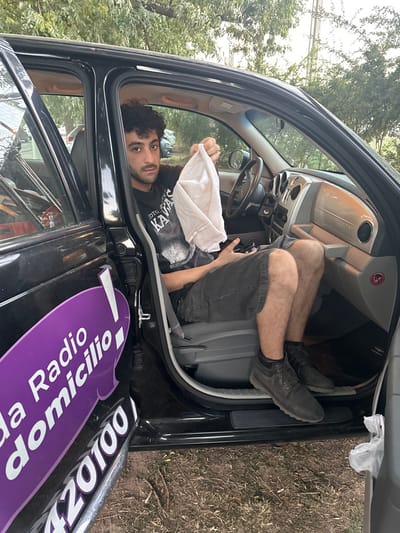 A man is lying in the driver's seat of a car, facing the camera. He holds a white cloth in his left hand. The car door is open, revealing a purple sign with white text, which appears to be a promotional sticker. The interior of the car is visible, including the dashboard and steering wheel. The man wears a black t-shirt with a white design, shorts, and boots. The background shows a glimpse of greenery and what appears to be a wooded area.