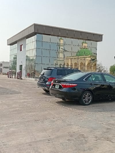 A two-story building with a facade composed entirely of reflective glass panels stands under a clear, bright sky in Kariya, Nigeria. The building features a flat, dark-trimmed roof. Its reflective surface prominently displays the reflection of a nearby mosque, which is characterized by a light-colored structure, multiple arched openings, a tall minaret, and a distinct green dome. In the foreground, parked on a light grey concrete surface, are two dark-colored vehicles: a rear-facing dark SUV (possibly a BMW X5) and a black Toyota Camry sedan, both positioned at an angle from the camera's perspective. Faint tire marks are visible on the concrete ground. Distant structures and minimal vegetation are discernible on the far left and right horizons. No individuals are visible.