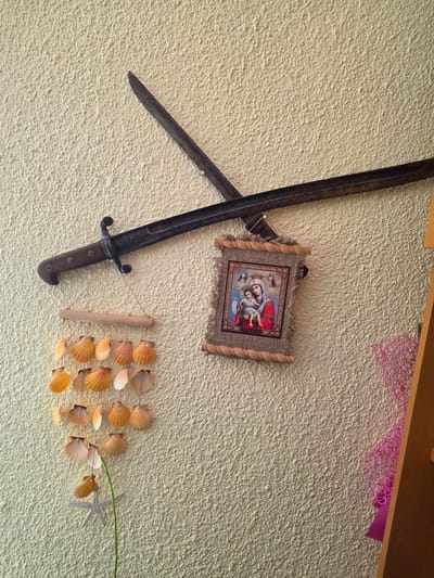 The image shows a textured, cream-colored wall with several decorative items mounted on it. The most prominent object is a long, dark metal bayonet with a wooden handle, angled diagonally across the upper portion of the frame. Below the bayonet, to the left, hangs a seashell wind chime made of numerous small, orange and white shells suspended from a wooden dowel. To the right of the wind chime and below the bayonet is a small, framed religious icon depicting the Virgin Mary and child, bordered with burlap and rope. Further to the right, a sliver of a wooden shelf or furniture is visible, along with a piece of pink, intricate netting. The wall appears to be indoors. There are no people or animals visible in the image. The scene suggests a personal decorative display within a residential or indoor setting, possibly in Targovishte, Bulgaria, given the prompt's contextual information.