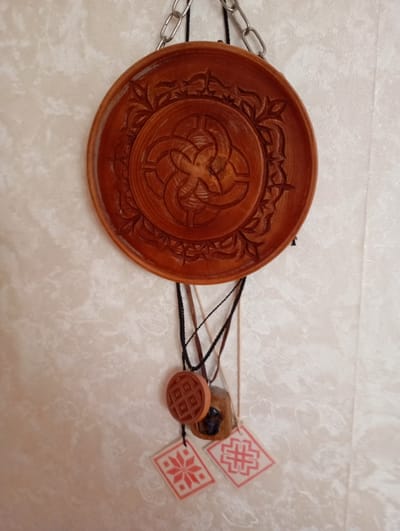 A circular, intricately carved wooden plate hangs from a metal chain against a textured, off-white wall. Attached to the plate by thin cords are three small decorative items. The first is a round, waffle-patterned wooden charm. Below it hangs a small, dark object, possibly wood or stone, with a rough texture. To the right of this is a square pendant with a red, geometric symbol on a white background. Another similar pendant, also with a red geometric symbol on white, is visible to the left of the first. The overall impression is that of a handcrafted decorative piece. This object is located in Kyshtym, Russia.