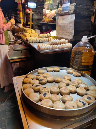 The image depicts a food stall operating in an evening market setting in Sa Pa, Vietnam, with a primary focus on various cakes and pastries. In the immediate foreground, a large, circular metal pan contains dozens of shallow-fried, golden-brown, flattened cakes. Some of these cakes are plain, while others are topped with black sesame seeds.
Directly behind this pan, stacked metal trays display rows of uniformly shaped, lighter-colored, oblong cakes. To the right, a large, transparent plastic bottle filled with yellow cooking oil stands upright.
On the left side of the frame, a person, dressed in a long white skirt and a pink top over a green garment, is partially visible from the waist down, with their left hand raised and appearing to handle a small item. An electronic weighing scale rests on a wooden counter positioned next to this individual. Further back, another person is discernible, seated behind a large, woven basket filled with dark, oval-shaped produce.
The stall's rear wall is constructed from dark, rough wooden planks. A bright, rectangular LED light fixture is mounted on the upper left section of this wall, providing illumination. A handwritten sign, affixed to the wooden surface, advertises "ONE REVIEW ON GOOGLE, ONE CAKE FREE," identifies the vendor as "Tiem nha Mong," and lists cake options including "Chestnut cake," "Green O Mix," and "Chestnut bean." A QR code is visible above this sign. The entire scene is artificially lit, suggesting an operation after sunset.