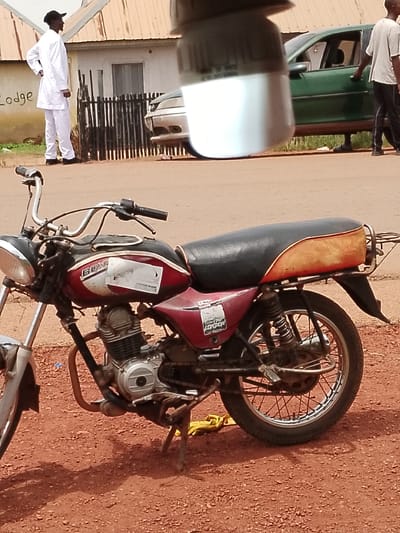 In Wumat, Nigeria, a red and black motorcycle is parked on a dirt road in front of a building. A man in a white tunic and pants, wearing a black cap, stands to the left of the motorcycle, facing away from the camera. A dark-colored car is parked behind a dark fence to the right of the building. The building has a light-colored corrugated metal roof and a light-colored wall with the word "LODGE" painted on it. The ground is covered in red dirt and small stones, with shadows cast from the motorcycle and an unknown object in the foreground.