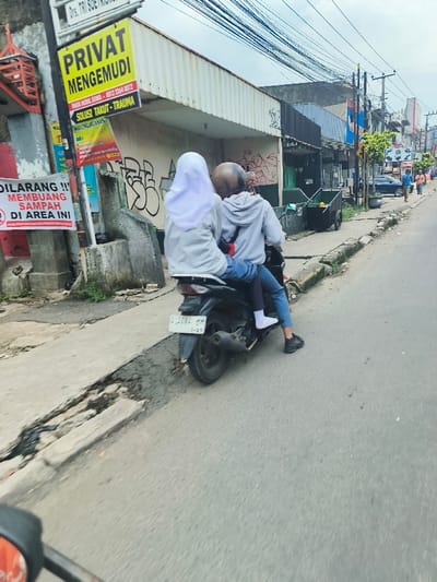 The image shows a black motorcycle parked on the side of a paved road in Cimahi, Indonesia (latitude: -6.878, longitude: 107.549). The motorcycle is facing towards the right of the image. Two people are riding the motorcycle. The person in the front is wearing a white hijab and a grey hoodie, while the person in the back is wearing a black helmet, a grey hoodie, and blue jeans. Both are wearing white socks and black shoes. The motorcycle is parked in front of a wall with graffiti on it. There are buildings on both sides of the road. There is a sign on a pole to the left of the motorcycle with Indonesian language and a sign with red text that says “DILARANG !! MEMBUANG SAMPAH DI AREA INI”. There are electrical wires hanging over the road and a trashcan on the right side of the motorcycle. There are other people walking on the sidewalk in the distance to the right of the image.  The scene is taken from the perspective of a person standing on the sidewalk.  The time of day is daytime, with an overcast sky.  The weather is cloudy.