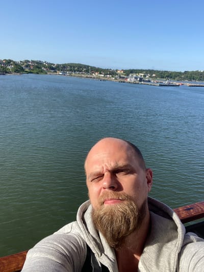 The image is a selfie taken by a man with a shaved head and a beard. He is wearing a grey hooded sweatshirt. He is standing on a wooden deck next to a body of water. In the background, across the water, is a city with buildings on a hill, as well as a harbor with several boats and ships. The sky is clear and blue. This scene is consistent with the waterfront area of Gothenburg, Sweden, potentially near a marina or harbor district.