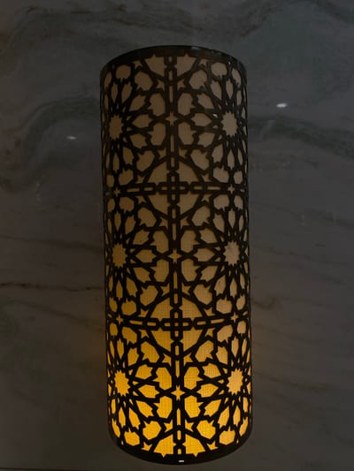 The image displays a close-up of a cylindrical light fixture, horizontally mounted against a dark, polished marble or stone wall. The light fixture is illuminated, emitting a warm, golden-yellow glow that highlights its intricate design. The outer shell of the fixture features a dark, laser-cut geometric pattern, characteristic of Islamic art and architecture. This pattern consists of repeating star-like and polygonal shapes, creating a complex tessellation. Behind this dark overlay, a lighter, possibly fabric or frosted glass, material diffuses the light, allowing the geometric cutouts to stand out in sharp contrast.
The background wall exhibits subtle, lighter veining typical of natural stone, reflecting a faint sheen from the light source. The overall lighting suggests an indoor setting, possibly an evening or night scene, where the decorative lamp provides ambient illumination. There are no people or specific activities depicted. The design of the light fixture strongly points to a Middle Eastern aesthetic, consistent with the provided location context of Doha, Qatar. No visible text is present in the image.