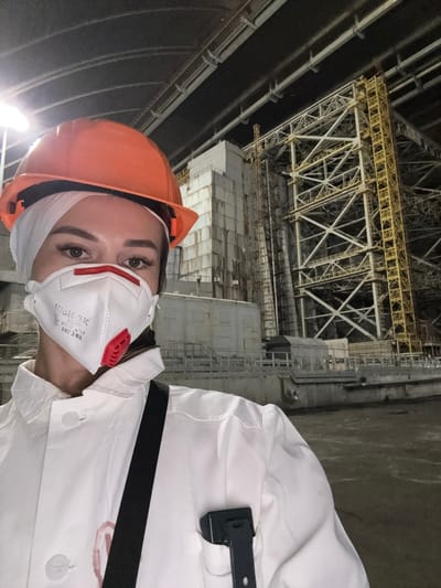 The image shows a woman wearing a white coat, an orange hard hat, and a white respirator mask. She is taking a selfie in what appears to be an industrial facility with large metal structures and concrete buildings in the background. The lighting is dim, with some artificial lights visible. The woman's face is partially obscured by the mask, but her eyes and eyebrows are visible. She has long, dark hair pulled back. A black strap is visible across her chest, and a black device is clipped to her coat pocket. The background suggests an area undergoing construction or renovation. This scene is located within the city of Kyiv, Ukraine.