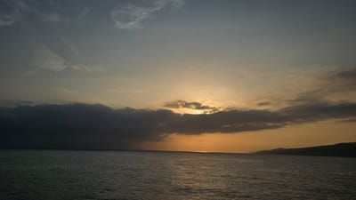 This media file captures a serene seascape at sunset, with the sun's rays breaking through a band of dark clouds, casting a warm orange glow over the horizon. The ocean is calm, reflecting the dramatic sky above. A distant coastline, likely Pasaia, Spain, is visible on the right, its silhouette softened by the fading light. The sky transitions from a deep, dusky blue at the top to a warm orange and yellow near the horizon, with wispy clouds scattered throughout. The overall mood is peaceful and contemplative, evoking the quiet beauty of nature as day turns to night. There is no visible text in the image.