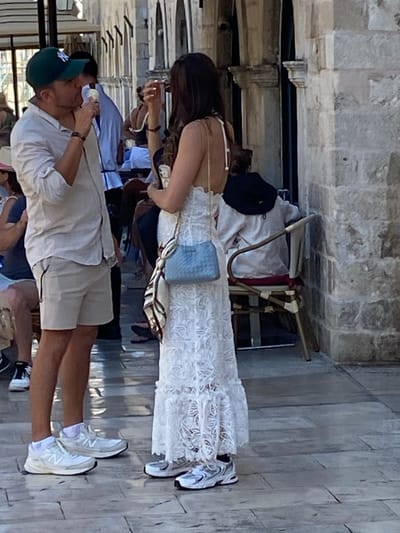 A man wearing a green New York Yankees baseball cap, a light-colored short-sleeved button-down shirt, and beige shorts is eating ice cream. He is standing next to a woman wearing a long white lace dress with a blue handbag slung over her shoulder and a patterned scarf tied to it. The woman is holding an item to her face. They are standing on a cobblestone street in Dubrovnik, Croatia, likely near the Stradun, a famous thoroughfare. In the background, other people are visible, some sitting at tables. The architecture suggests historic buildings with stone facades. The lighting indicates it is daytime.