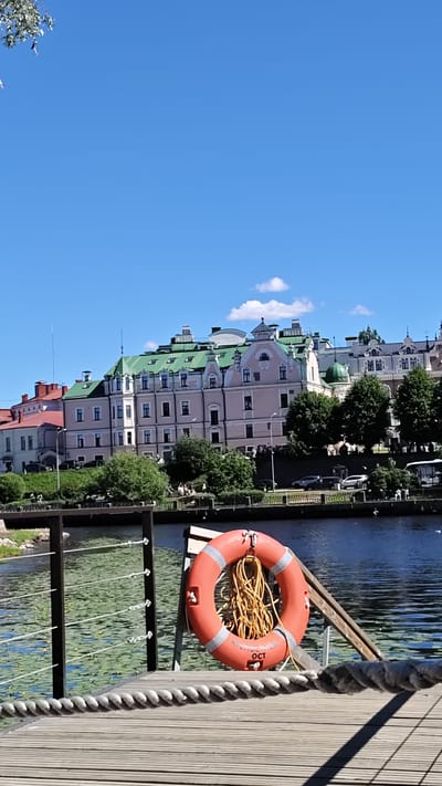 The image captures a waterfront scene in Vyborg, Russia, with a focus on a wooden pier in the foreground and historic buildings in the background. A bright orange life preserver with a coiled yellow rope hangs on a wooden stand to the right of the pier railing. The pier itself is made of weathered wooden planks and is bordered by a sturdy railing with rope accents. Beyond the pier, a calm body of water, possibly a canal or bay, reflects the bright blue sky and is dotted with green lily pads.
In the middle ground, a row of old buildings with distinct green roofs and pale pink facades lines the opposite bank of the water. These buildings feature numerous windows and architectural details characteristic of historical European architecture. Trees with lush green foliage are interspersed between the buildings and along the waterfront, adding to the scenic quality of the urban landscape. A road or pathway runs in front of the buildings, with a few parked cars visible.
The sky is a clear, vibrant blue, with a few wispy white clouds scattered across the upper portion of the image. The overall impression is one of a sunny day in a historic city, with the waterfront serving as a central element of the view. The image appears to be taken from a slightly elevated position, looking across the water towards the city.