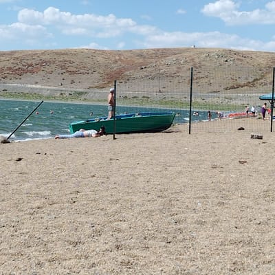 A beach scene is depicted with a body of water and a dry, hilly landscape in the background. In the foreground, a sandy beach is visible. A person is lying on their stomach on the sand, facing away from the viewer, with their legs extended. They are wearing a light-colored t-shirt and blue jeans. Behind this person is a green boat with a blue trim, resting on the sand. A man is standing in the boat, wearing dark shorts and a grey cap, facing away from the camera. A metal fence runs horizontally across the middle of the image, separating the foreground from the background. In the water, several people are visible, some swimming and others near the shore. The water appears choppy, indicating wind. In the distance, the landscape consists of undulating, dry hills with sparse vegetation and scattered rocks. The sky is blue with scattered white clouds. The overall setting suggests a recreational area by a body of water.