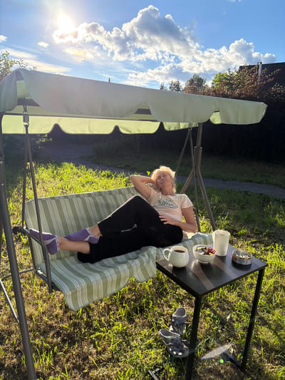 A woman is relaxing on a green and white striped swing seat in a backyard in Steklyannyi, Russia. She is wearing a light-colored t-shirt, black pants, and purple socks. Her head is propped up on her hands, and her eyes are closed. A small black table is next to the swing, with two mugs of coffee, a bowl of candy, and a small tin can on it. A pair of grey Crocs are on the ground next to the table. The sun is shining brightly, casting long shadows across the grass. The sky is blue with scattered white clouds. In the background, there are trees and a house.