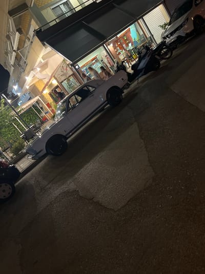 The image depicts a street scene at night in Νέα Φλογητά, Greece. A white classic car, possibly a Ford Mustang, is parked on the asphalt road, angled towards the viewer. The car has a blue stripe along its side and black wheels. In the background, there is a building with multiple levels, featuring balconies and awnings. One section of the building appears to be a shop or restaurant with illuminated windows and signage that includes Greek text. A white SUV and a dark-colored scooter are parked behind the classic car, further down the street. Several people are visible inside the illuminated establishment, and some are standing outside near the scooter. The street appears to be wet, reflecting the artificial lights from the buildings and possibly from the streetlights. Greenery, such as ivy or vines, is visible on the side of one of the buildings. The overall atmosphere suggests a commercial area at night.