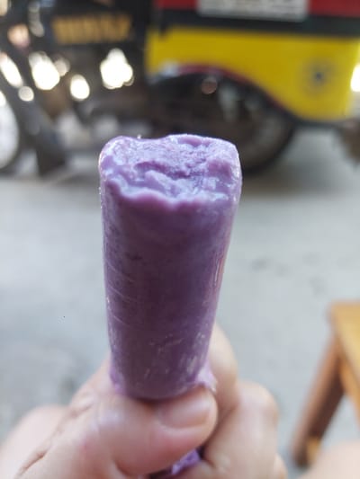 A close-up, slightly out-of-focus shot shows a hand holding a purple popsicle wrapped in clear plastic. The popsicle has a bumpy, textured surface and a vibrant purple color. In the blurred background, a yellow and black object, possibly a vehicle or cart, is visible. The scene appears to be outdoors, with a light-colored ground and hints of other objects in the periphery. The overall impression is casual, likely taken in a public or semi-public space in Samal, Philippines.