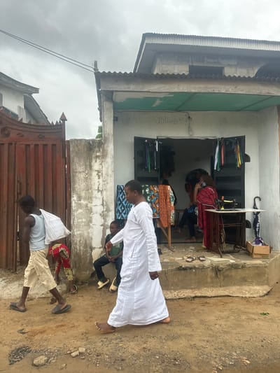 The image depicts an outdoor scene in Ajara, Nigeria, with a focus on a small tailor shop. The shop appears to be a modest building with a corrugated metal roof and a light-colored facade. Inside, a woman in a red and black patterned dress is seated at a sewing machine, actively engaged in tailoring. Various fabrics, some folded and stacked on shelves, and a selection of colorful umbrellas are visible within the shop.
Several people are present in front of and around the shop. Two individuals, both appearing to be men, are standing near the entrance. One, wearing a white robe, is facing away from the camera and looking down, possibly at the ground or at someone. Another person, partially visible in a green shirt, is seated nearby. Further down, a young boy in a light blue shirt and tan shorts is lying on a metal gate, appearing to be resting or waiting. Another person in a red and yellow patterned skirt and white top is also visible near the shop.
The ground is uneven and appears to be dirt or sand, with some scattered gravel. The weather seems overcast, suggesting a cloudy or possibly rainy day. The overall atmosphere is casual and suggests everyday life and commerce in a Nigerian community. There is some visible text on the wall of the shop: "08124623919". The image is oriented vertically.
