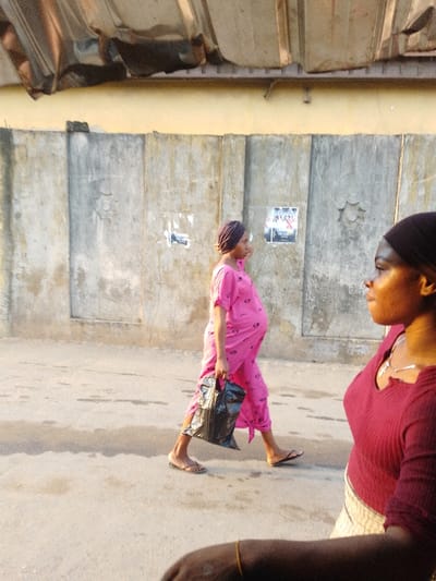 A pregnant woman in a pink robe and headscarf walks on a street holding a black bag. A second woman wearing a maroon top and head covering stands in the foreground, partially obscuring the view. The background features a concrete wall with posters or markings. The scene appears to be outdoors during daylight.